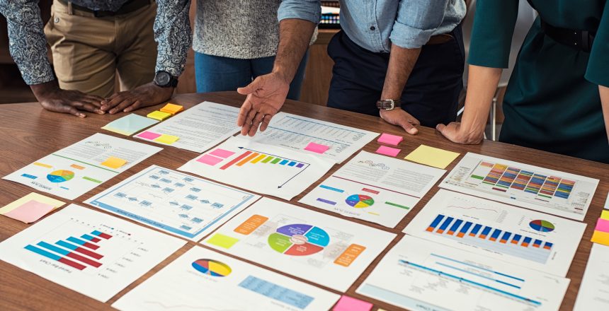 Financial and business documents on table with multiethnic hands working on it. Latin business manager with colleagues working on new startup project. Closeup business man and businesswoman hands understanding pie and bar graphs during meeting.
